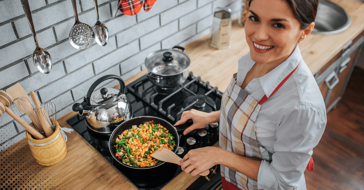 Woman smiling while cooking mixed vegetables on a stovetop in a cozy kitchen.